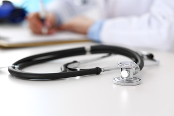 Closeup of stethoscope. Female doctor fills up medical form while sitting at the desk in hospital. Healthcare, workplace and cardiology in medicine concept