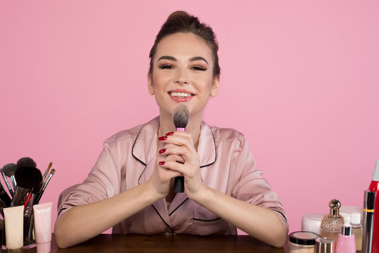Portrait of cheerful girl cosmetologist is sitting at dressing table and holding professional makeup brush. She is looking at camera with joy. Isolated on pink background