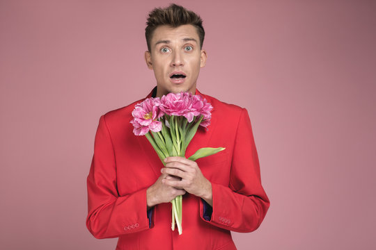Portrait of young guy with shocked expression keeping rose flowers with both hands. He is wearing red jacket. Isolated on background