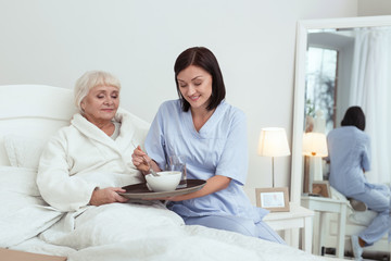 Time to eat. Merry nurse getting breakfast to elder woman who lying in bed