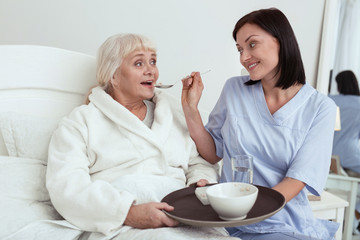 Meal for you. Jolly nurse feeding to elder woman who lying in bed