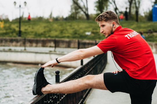 Handsome sporty guy is streching in a green beautiful park and is getting ready for jogging while is listening music.