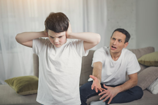 I do not want to hear you. Sad boy is closing his ears while his dad is shouting at him. Angry man is sitting on couch on background