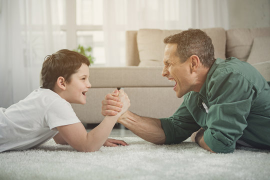 Joyful man and boy are competing in arm wrestling while lying on carpet in living room. Child is looking at dad and smiling