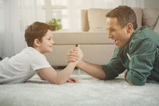 Lets see who is stronger. Cheerful father and son are doing arm-wrestling. They are lying on floor and laughing