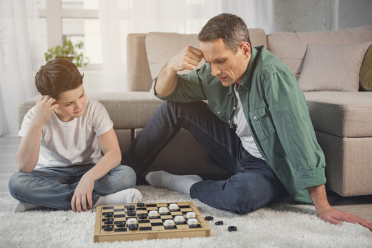 Pensive father and son are playing checkers together in living room. They are looking at board and thinking