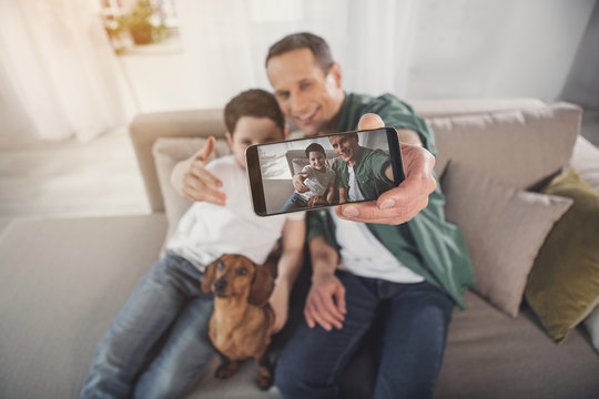 Happy man is making selfie with his son and dog on smartphone. They are embracing and smiling while sitting on sofa. Focus on gadget screen