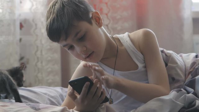 boy lies in bed in his bedroom in the morning and uses a smartphone.