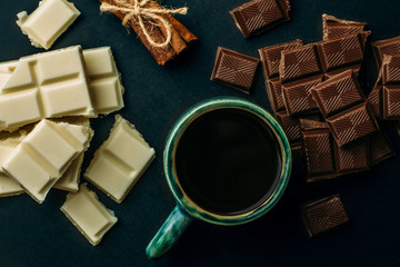 Cup of coffee, broken white and black chocolate bars and stack of cinnamon on black table, top view. Tasty energy calorie food