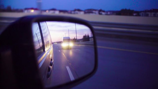 Rear view mirror in a moving car at night. It reflects traffic on the road, forest and cloudy sky.