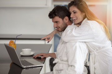 young couple in bathrobe at home having coffee in the kitchen and working on laptop computer