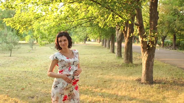 Happy pregnant girl in beautiful cocktail dress walking in the Sunny summer Park at sunset. Pregnant woman gently stroking her tummy in the Park at sunset. Slow motion.