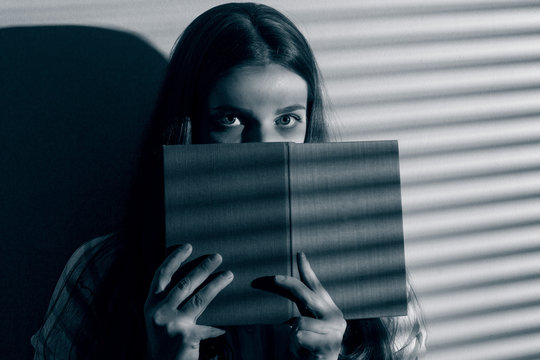 A young woman scaredly hides behind the book, which reads the light of the night.