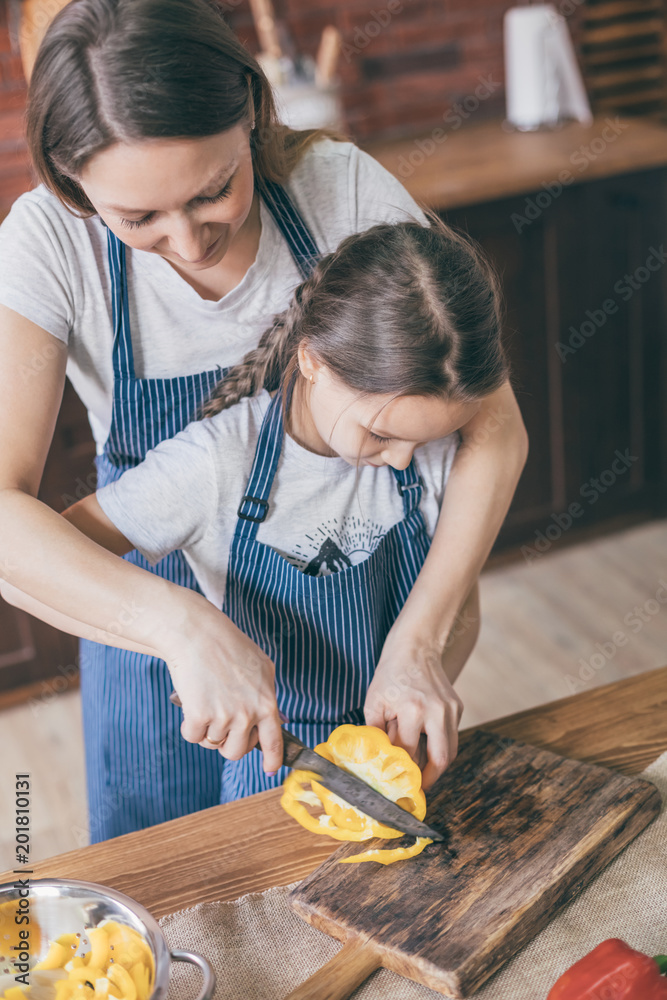 Poster adult woman standing and helping to daughter to cut yellow pepper on kitchen at home.