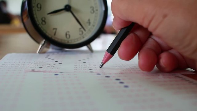 Asian Students taking optical form of standardized exams near Alarm clock with hand holding yellow pen for final examination in secondary school, college university classroom, Education concept