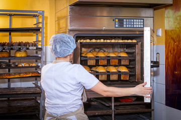 A female baker in white uniform and a beige apron bakes bread and takes cheese buns from an...