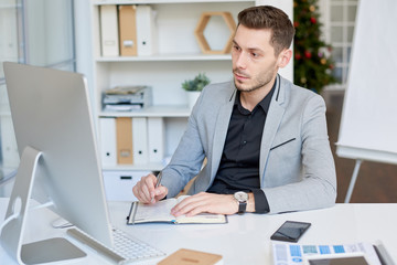 Fototapeta na wymiar Portrait shot of handsome entrepreneur with stylish haircut making video call to business partner and taking necessary notes while sitting at modern office
