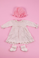 Cute girl clothes. A white little dress and a knitted hat. Pink background.