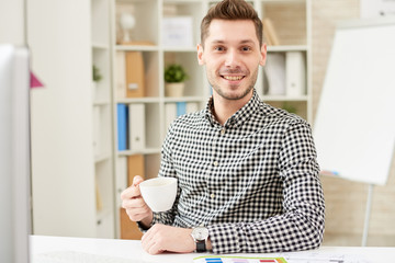 Waist-up portrait of handsome young manager wearing checked shirt looking at camera with wide smile while sitting at office desk and drinking fragrant coffee