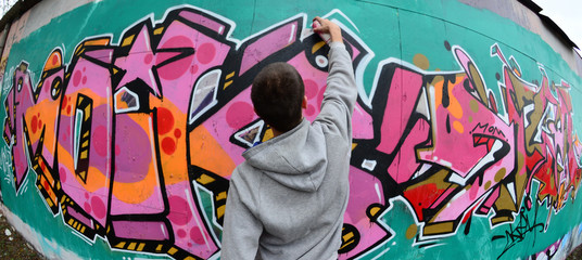A young guy in a gray hoodie paints graffiti in pink and green colors on a wall in rainy weather....