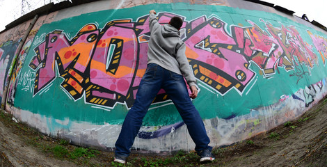 A young guy in a gray hoodie paints graffiti in pink and green colors on a wall in rainy weather....