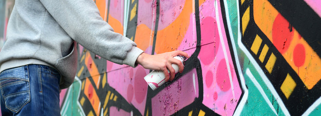 Hand of a young guy in a gray hoodie paints graffiti in pink and green colors on a wall in rainy weather