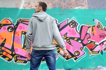 A young graffiti artist in a gray hoodie looks at the wall with his graffiti in pink and green colors on a wall in rainy weather. Street art concept