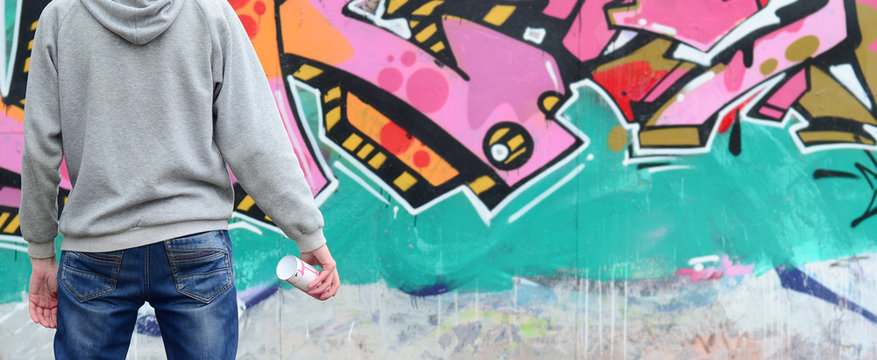 A young graffiti artist in a gray hoodie looks at the wall with his graffiti in pink and green colors on a wall in rainy weather. Street art concept