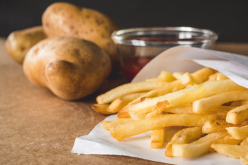 Close up of French fries with ketchup and raw potatoes on wooden table.