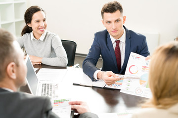 Positive successful male manager presenting statistical data and pointing at pie chart while explaining charts to colleagues at business meeting