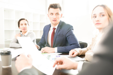 Serious curious company employees sitting in a row at table and listening to boss who explaining documentary at staff meeting