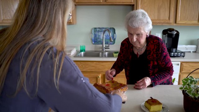 Mature woman helps elderly woman put pineapple upside down cake onto a plate.