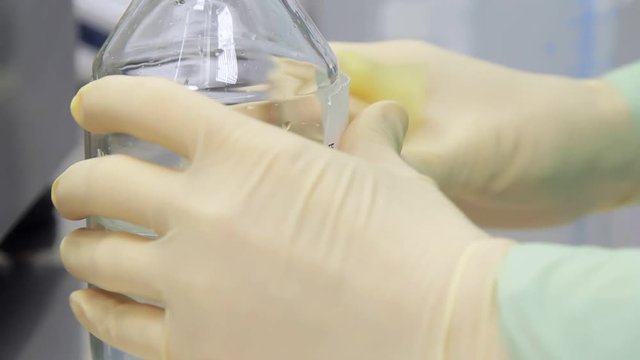 Hand in gloves stick water bottle. Close up of laboratory worker sticking sticker at bottle with transparent liquid. Laboratory research