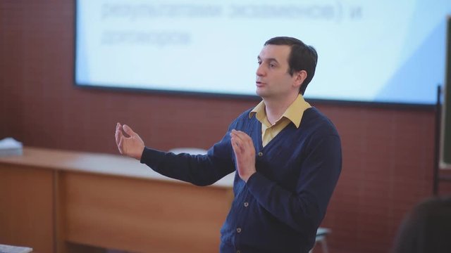 Teacher at the university gives a lecture. Male professor is teaching students. Male teacher is giving a lecture at the university. Professor stands on background of presentation from the projector