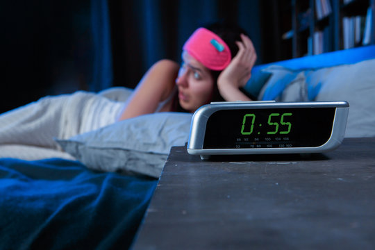 Image of woman with insomnia with pink bandage for eyes lying on bed next to clock