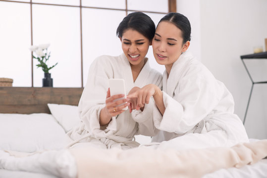 Not the best choice. Nice positive attractive women sitting together on the bed while looking at the photos on their smartphone