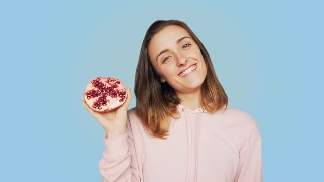 Cute, pretty and adorable young woman, smiles and laughs, excited and amused, happy with real emotions, twists and bops her head rhythmically with music, holds fresh juicy red pomegranate