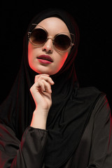 Portrait of beautiful stylish young muslim woman wearing black hijab and sunglasses as modern eastern fashion concept posing on black background