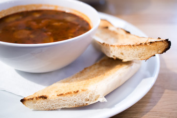 Tasty soup on white plate with pastry detail, Red soup with fried baguette, Dark universal soup, Vegetable soup with baguette on wooden table