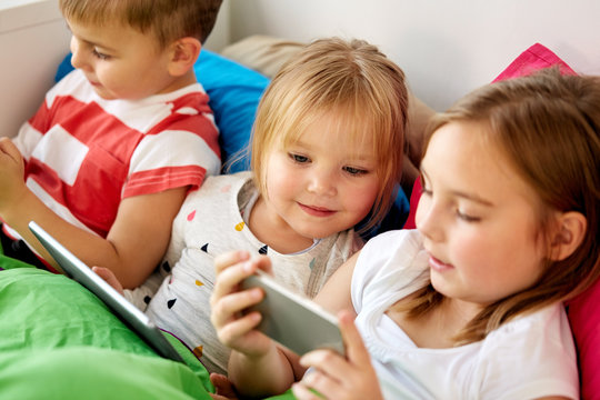 childhood, technology and family concept - happy little kids with smartphone and tablet pc computer in bed at home