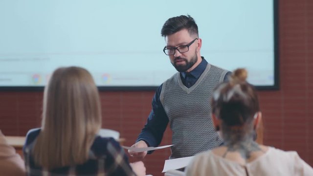 A young university teacher distributes to students the sheets of paper with the results of a test. Lecture at the University: students and a young professor with a beard.