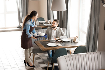 Smiling content beautiful waitress holding tray with tea and giving order to customer sitting at table and talking to her in modern cafe