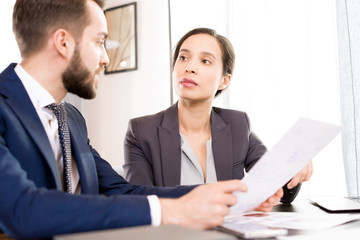 Serious pensive attractive Hispanic businesswoman listening to colleagues offer while they doing paperwork and analyzing data together in office