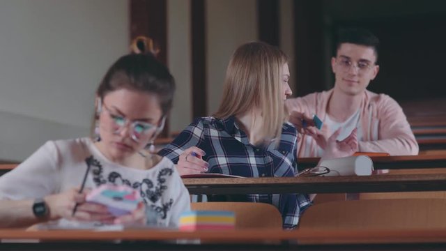 A female student unfolds a note, which was given by a classmate and then smiles. Cheat sheet, love note. Group of three modern students in lecture theater: guy and two female students.