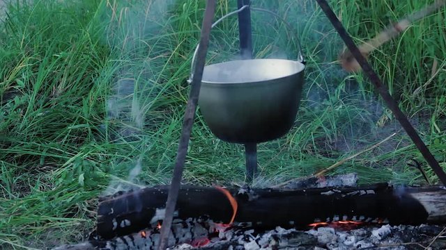 Bowler at the stake The fisherman removes scaling with a wooden spoon in a bowler hat suspended on a tripod over a campfire close up