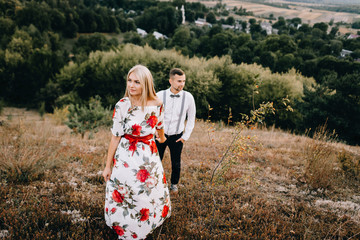 Attractive young loving couple of man in white shirt and with suspenders and gentle girl in dress with red flowers are walking on sunny mountain background in sunset. Hold hands