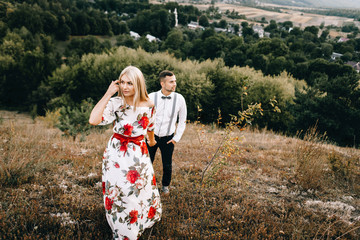 Attractive young loving couple of man in white shirt and with suspenders and gentle girl in dress with red flowers are walking on sunny mountain background in sunset. Hold hands