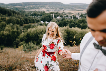 Attractive young loving couple of man in white shirt and with suspenders and gentle girl in dress with red flowers are walking on sunny mountain background in sunset. Hold hands