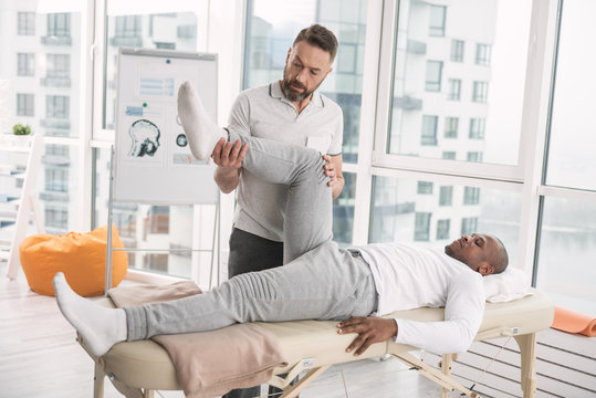 Physical therapist. Serious nice bearded doctor holding and looking at the patients leg while doing the therapy
