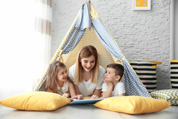 Nanny and little children reading book in tent at home - obrazy, fototapety, plakaty